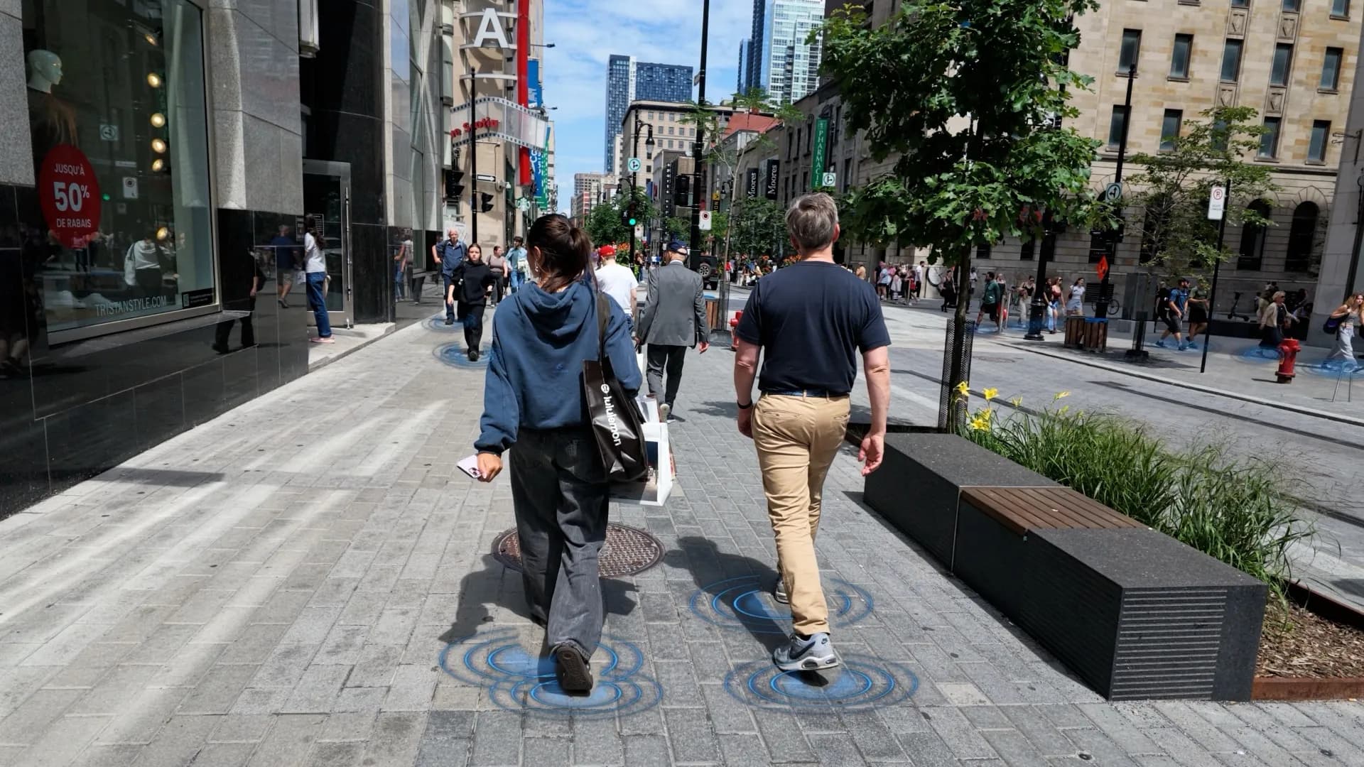 Two people walk away up a Montreal shopping street in summer, storefronts and street trees on either side, rings of blue light spreading on the paving under each step